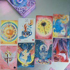 May include: A collection of Cherry Dreamwoven Lenormand oracle cards spread out, featuring vibrant watercolour illustrations including a sun, a moon, a key, an anchor, a market scene, a bed, a cross with wings, and a cup with a plant.