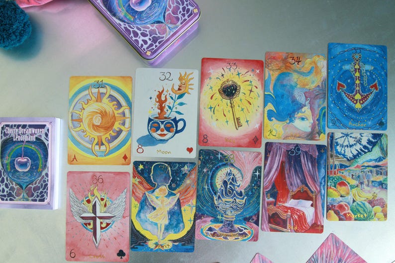May include: A collection of Cherry Dreamwoven Lenormand oracle cards spread out, featuring vibrant watercolour illustrations including a sun, a moon, a key, an anchor, a market scene, a bed, a cross with wings, and a cup with a plant.