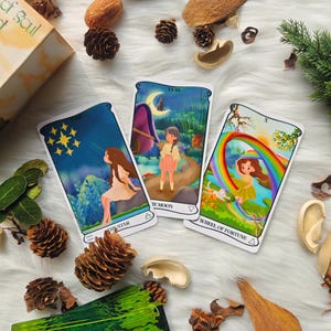 May include: Three tarot cards are displayed, featuring whimsical illustrations. The cards are labeled "The Star," "The Moon," and "Wheel of Fortune." Each card depicts a scene with a person and colorful imagery, including a rainbow and a starry night sky. The cards are on a white, fluffy surface.