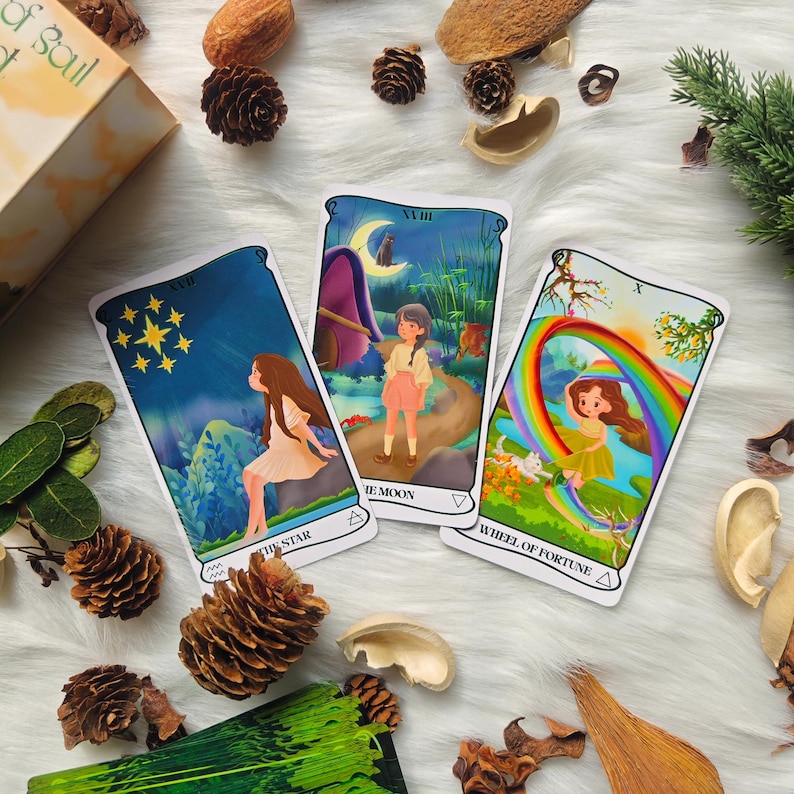 May include: Three tarot cards are displayed, featuring whimsical illustrations. The cards are labeled "The Star," "The Moon," and "Wheel of Fortune." Each card depicts a scene with a person and colorful imagery, including a rainbow and a starry night sky. The cards are on a white, fluffy surface.
