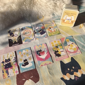K&ouml;nnte beinhalten: Ein Satz Mystic Meows Tarotkarten mit skurrilen Katzenillustrationen. Die Karten werden mit einer gelben Blechdose pr&auml;sentiert, und die Karten tragen Titel wie "Gericht", "Die Welt" und "Sieben der Kelche". Die Farbpalette umfasst Pastellt&ouml;ne und weiche Farbt&ouml;ne.