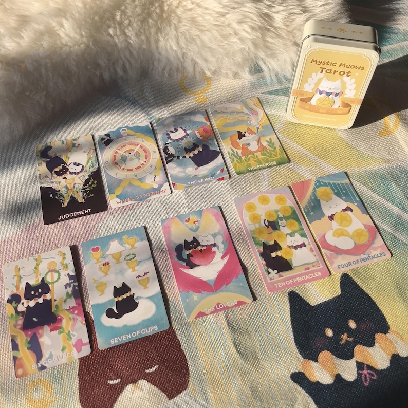 K&ouml;nnte beinhalten: Ein Satz Mystic Meows Tarotkarten mit skurrilen Katzenillustrationen. Die Karten werden mit einer gelben Blechdose pr&auml;sentiert, und die Karten tragen Titel wie "Gericht", "Die Welt" und "Sieben der Kelche". Die Farbpalette umfasst Pastellt&ouml;ne und weiche Farbt&ouml;ne.