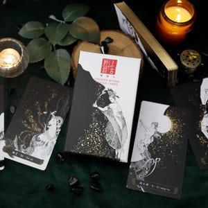 May include: A deck of "Ancient Myths Oracle Cards" with a white and black box featuring an illustration of a person. Several black and white oracle cards with gold accents are scattered around the box. A lit candle and other decorative items are also present.