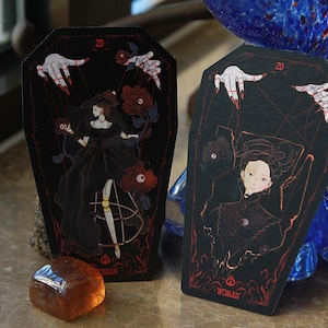 May include: Two coffin-shaped tarot cards with black backgrounds and red line art. The cards feature illustrations of a figure in a black dress, red roses, and pale hands with red fingernails. Each card displays the number 29 and the word WOMAN.