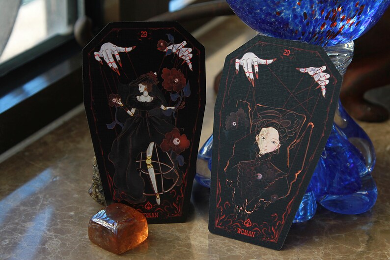May include: Two coffin-shaped tarot cards with black backgrounds and red line art. The cards feature illustrations of a figure in a black dress, red roses, and pale hands with red fingernails. Each card displays the number 29 and the word WOMAN.
