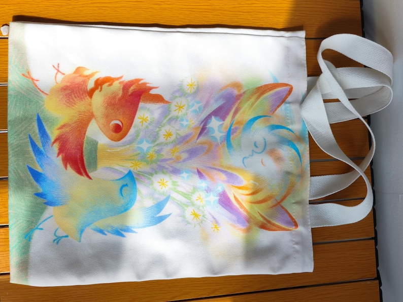 May include: White tote bag with a colorful illustration of birds and floral elements. The artwork features a bird with orange and yellow feathers, and another with blue and yellow feathers. A white strap is attached to the bag.