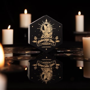 May include: A hexagonal black box featuring gold foil illustrations of a raven and sunburst, with the text Shadow Night Raven Lenormand printed on the front. The box is reflected on a dark surface, surrounded by several out-of-focus white candles.