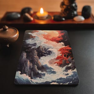 May include: A rectangular print featuring a landscape illustration with jagged dark grey mountains, swirling white and blue ocean waves, a vibrant red tree, and a large glowing orange moon set against a dark sky with clouds.
