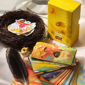 May include: A yellow box with the text "suno with the Sun" next to a deck of colorful cards with illustrations. A small bird figurine, a black feather, and a nest with stickers are also present.