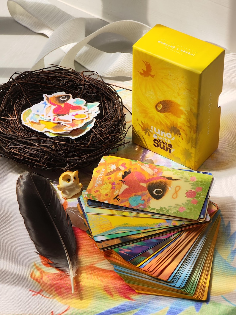 May include: A yellow box with the text "suno with the Sun" next to a deck of colorful cards with illustrations. A small bird figurine, a black feather, and a nest with stickers are also present.