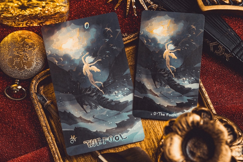 Op de afbeelding: Twee tarotkaarten met een hemelse illustratie van een gevleugelde figuur. De kaarten zijn donkerblauw met de woorden "THE FOOL" en "0. The Fool" erop gedrukt. De achtergrond is een donkere, bewolkte lucht met helder licht.