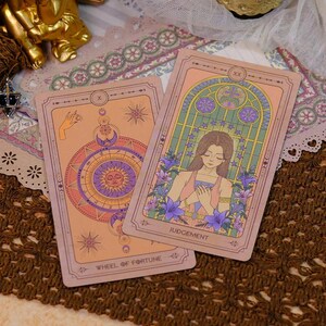 K&ouml;nnte beinhalten: Zwei Tarotkarten mit Vintage-&Auml;sthetik. Eine Karte zeigt ein Sonnen- und Monddesign mit dem Text "WHEEL OF FORTUNE". Die andere Karte zeigt eine Frau, umgeben von Blumen, mit dem Wort "JUDGEMENT". Die Karten haben ein rosa-violettes Farbschema.