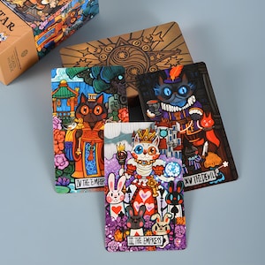 May include: A tarot card deck featuring colorful illustrations of cats in various roles, including "The Emperor" and "The Empress." The deck includes a wooden box with a detailed design and several individual cards with unique artwork.