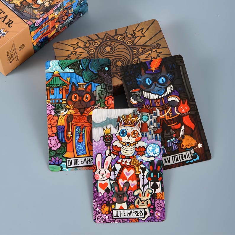 May include: A tarot card deck featuring colorful illustrations of cats in various roles, including "The Emperor" and "The Empress." The deck includes a wooden box with a detailed design and several individual cards with unique artwork.