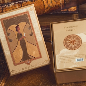May include: Two rectangular boxes with gold-colored frames. One box has an illustration of a woman in a dress, and the text "LENORMAND". The other box has the text "PENTHOUSE BANQUET" and a circular design. The boxes are on a gold-colored surface.