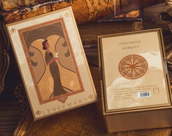 May include: Two rectangular boxes with gold-colored frames. One box has an illustration of a woman in a dress, and the text "LENORMAND". The other box has the text "PENTHOUSE BANQUET" and a circular design. The boxes are on a gold-colored surface.