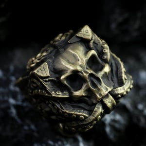 Può includere: Un anello dettagliato color bronzo con un design a teschio. Il teschio è intagliato in modo intricato con orbite oculari, cavità nasale e denti visibili. L'anello ha una fascia ornata e strutturata con elementi decorativi aggiuntivi.
