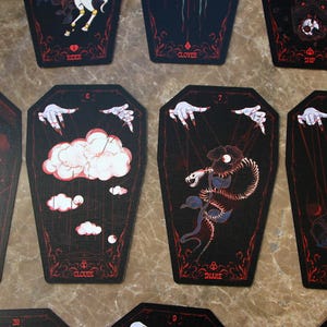May include: A set of coffin-shaped oracle cards with black backgrounds and red line art. The cards feature illustrations of hands with strings, clouds, a snake, and a coffin, with text labels including Clouds, Snake, and Coffin printed below.