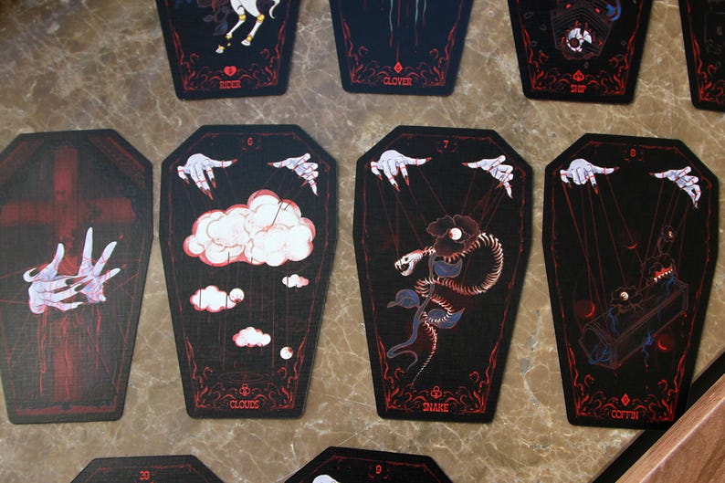 May include: A set of coffin-shaped oracle cards with black backgrounds and red line art. The cards feature illustrations of hands with strings, clouds, a snake, and a coffin, with text labels including Clouds, Snake, and Coffin printed below.