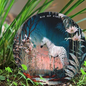 May include: Round decorative art piece featuring a zebra standing in a forest scene with the text "SIX OF FIRE". The artwork has a dark blue background with pink and white flowers and foliage.