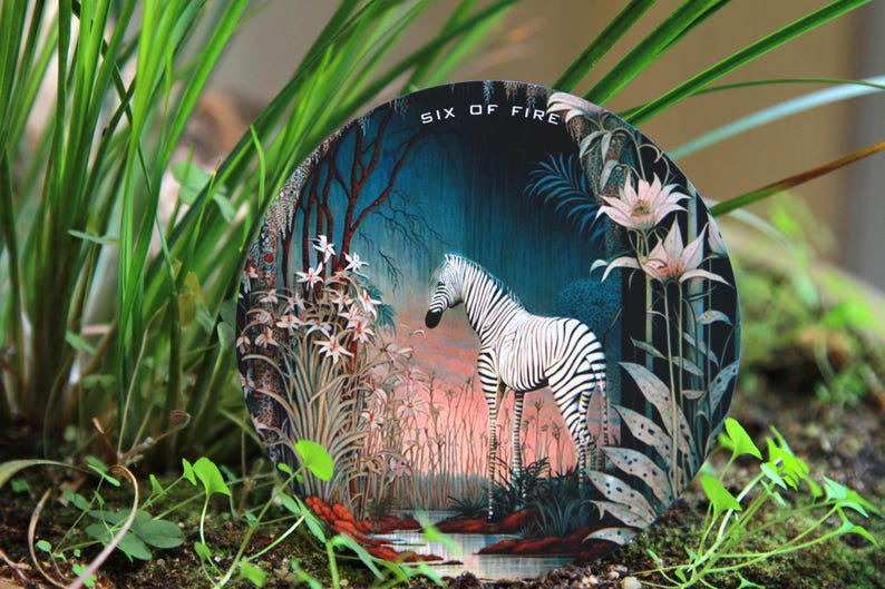 May include: Round decorative art piece featuring a zebra standing in a forest scene with the text "SIX OF FIRE". The artwork has a dark blue background with pink and white flowers and foliage.