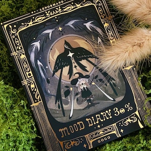 May include: A black and gold illustrated mood diary with the text "LENORMAND" and "MOOD DIARY 30%". The cover features a bird and floral design with a dark background and gold accents. The diary is set against a backdrop of green moss and dried grass.