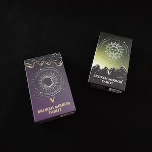 May include: Two rectangular tarot card decks in boxes. The left box is purple with a gold sun and moon graphic and the text BROKEN MIRROR TAROT. The right box is green with a gold sun graphic, clouds, and the text BROKEN MIRROR TAROT.