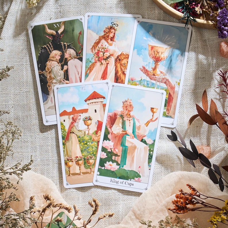 May include: A collection of tarot cards featuring watercolor illustrations. The cards depict various scenes, including figures with a lion, a devil, and a king holding a cup. The "King of Cups" card is visible. The cards are arranged on a neutral-toned surface.