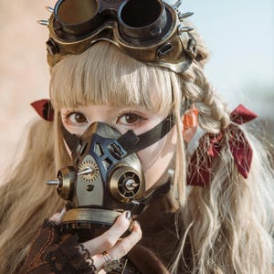 May include: A woman wearing a steampunk-style gas mask with a gold and black finish. The mask has a gear design and spikes. She is also wearing goggles with a gold frame and black lenses.
