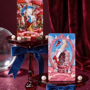May include: Two rectangular tarot cards featuring Alice in Wonderland themes. The left card, titled 12. The Hanged Man, shows a girl hanging upside down in a mushroom forest. The right card, 0. The Fool, depicts legs in red shoes and white socks.