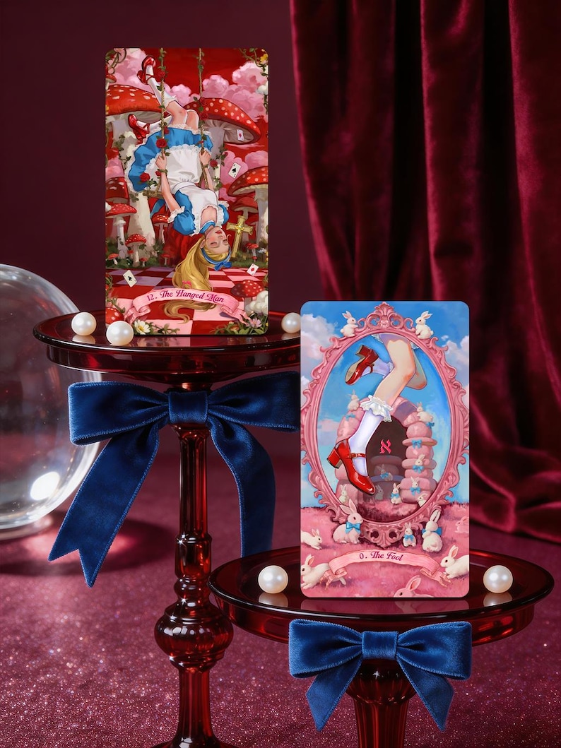 May include: Two rectangular tarot cards featuring Alice in Wonderland themes. The left card, titled 12. The Hanged Man, shows a girl hanging upside down in a mushroom forest. The right card, 0. The Fool, depicts legs in red shoes and white socks.