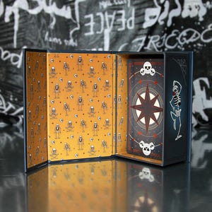 May include: Open tarot card box with a navy blue exterior and a mustard yellow interior patterned with skeleton illustrations. The box contains a deck of cards with a compass rose design and skull accents.