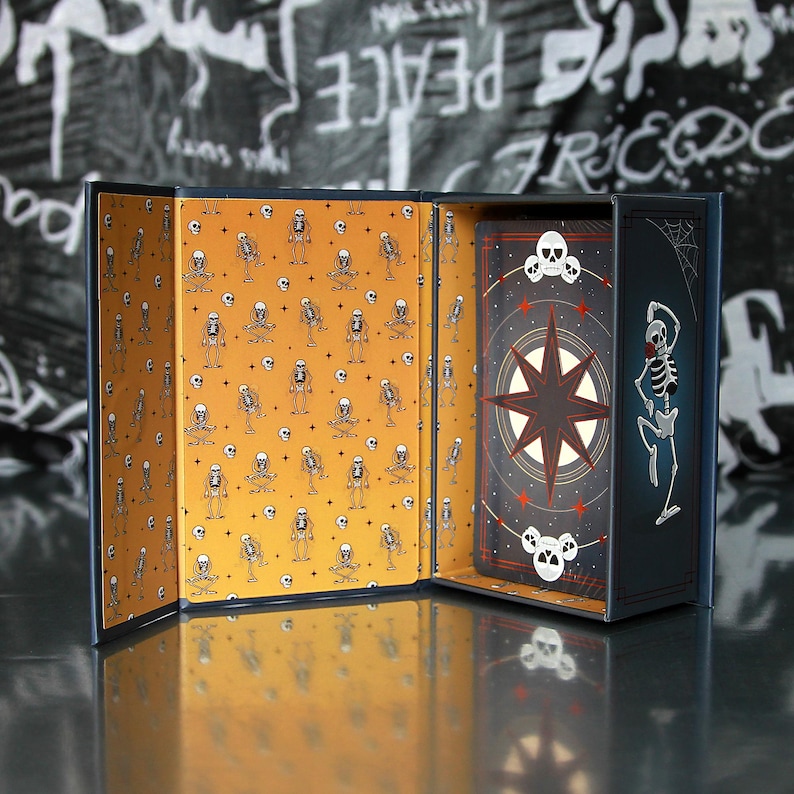 May include: Open tarot card box with a navy blue exterior and a mustard yellow interior patterned with skeleton illustrations. The box contains a deck of cards with a compass rose design and skull accents.