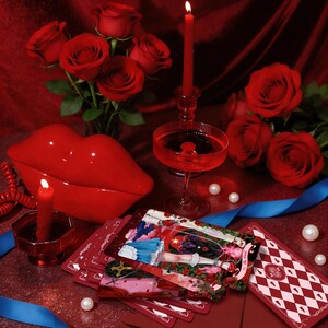 May include: A red ceramic lip-shaped container, a deck of tarot cards featuring a character in a blue dress, red roses, two red candles in glass holders, a red envelope with a wax seal, blue ribbons, and scattered white pearls on a red surface.