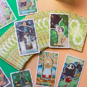 May include: A collection of tarot cards featuring whimsical illustrations and titles like "The Hermit" and "The Sun." The cards have a white border and are set against a floral green background. The artwork includes cartoon-style characters and symbols.