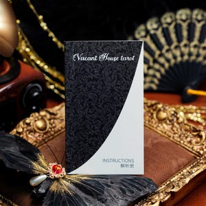 May include: A rectangular instruction booklet titled Vacant House tarot with a black cover featuring a white floral pattern on the left and a solid white section on the right. The text INSTRUCTIONS and Chinese characters are printed in black.
