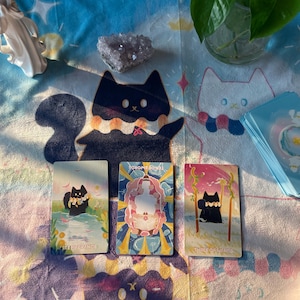 K&ouml;nnte beinhalten: Ein Stillleben mit Tarotkarten mit Katzenillustrationen, einem Kristall und einer Pflanze in einer Glasvase. Die Karten liegen auf einer gemusterten Decke mit Katzendesigns. Die Karten tragen die Aufschriften "Temperance", "Ten of Swords" und "Three of Wands".