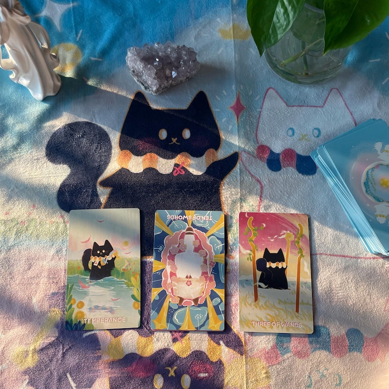 K&ouml;nnte beinhalten: Ein Stillleben mit Tarotkarten mit Katzenillustrationen, einem Kristall und einer Pflanze in einer Glasvase. Die Karten liegen auf einer gemusterten Decke mit Katzendesigns. Die Karten tragen die Aufschriften "Temperance", "Ten of Swords" und "Three of Wands".