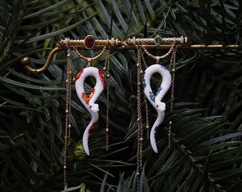May include: A pair of snake-shaped earrings. The white snakes have colorful patterns and hang from a gold-colored bar adorned with gemstones and chains. The background is a blurred view of green foliage.