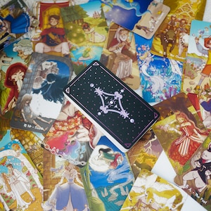 May include: A collection of tarot cards spread out on a table. The cards feature colorful illustrations and intricate designs. One card is face up, showing a black background with a white, star-shaped design. The card is labeled "Death".