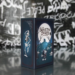 May include: A dark blue box with the text "The Skeleton Dance Arcana" and a cartoon skeleton dancing on one side. The other side features a cartoon character in a graveyard scene with a full moon. A spider web is in the corner.