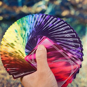 May include: A deck of playing cards fanned out in a circular shape, displaying a vibrant rainbow gradient of colours from red and orange to green, blue, and deep purple. Each card features a metallic, reflective finish and a white geometric line pattern.