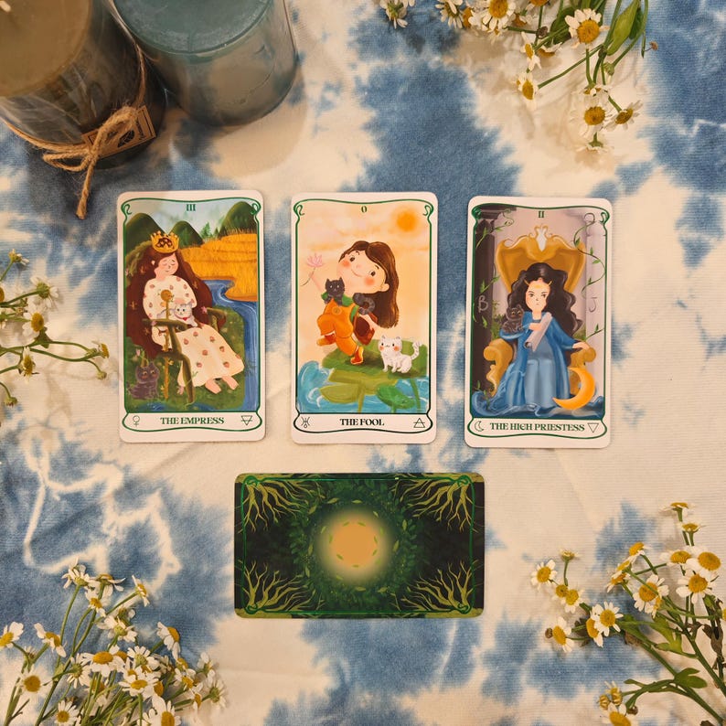 May include: Three tarot cards are displayed, featuring illustrations of The Empress, The Fool, and The High Priestess. The cards are set against a blue and white tie-dye background, with a deck of cards and flowers. The Empress card shows a woman with a crown.