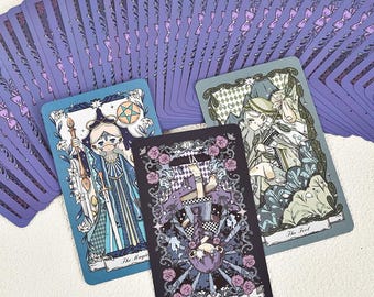 May include: A deck of tarot cards with purple backs featuring a repeating bow pattern. Three cards are fanned out: The Magician in blue, The Hanged Man in dark purple with pink roses, and The Fool in muted green, each with detailed illustrations.