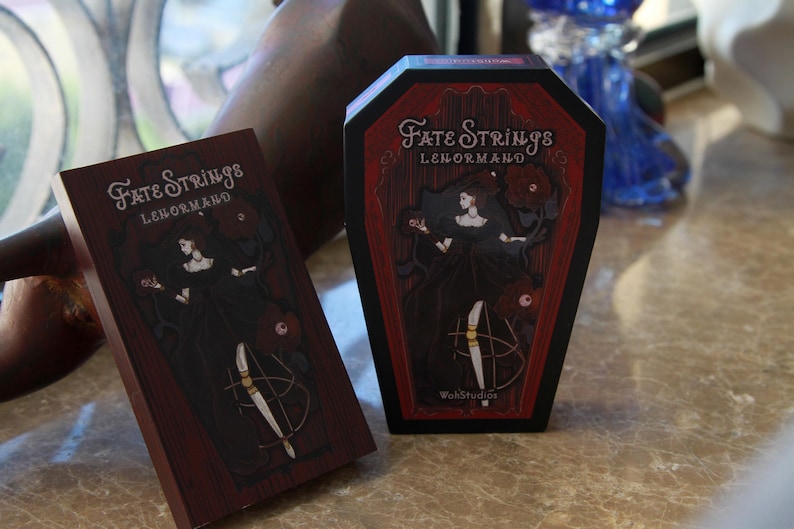 May include: A rectangular deck box and a coffin-shaped box for the Fate Strings Lenormand card set. Both feature a dark red and black illustration of a figure in a black dress with red flowers and the text Fate Strings Lenormand.