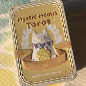 K&ouml;nnte beinhalten: Gelbe Tarotkartenbox mit den Worten "Mystic Meows Tarot" in goldfarbenen Buchstaben. Die Box zeigt eine wei&szlig;e Katze mit blauem und rosa Kragen, die ein goldenes Pentagramm h&auml;lt. Dekorative Elemente sind Katzen-Silhouetten und ein Band.