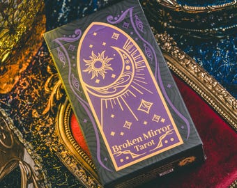 May include: A tarot card box with a purple and gold design. The box features a sun and moon illustration, with the words "Broken Mirror Tarot" in gold lettering. The box is on a red velvet surface.