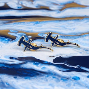 May include: Two gold-toned hammerhead shark pins with blue enamel. The pins are on a blue and gold marbled surface.