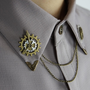 Puede incluir: Un broche de cuello dorado con un diseño de engranaje y una cadena. El broche está sujeto a una camisa gris.