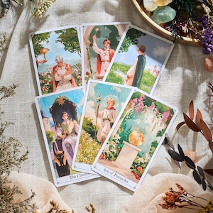 May include: A collection of tarot cards featuring detailed illustrations with a vintage aesthetic. The cards depict figures in classical attire and scenes with floral and architectural elements. The text "The Chariot" and "Ace of Pentacles" are visible.